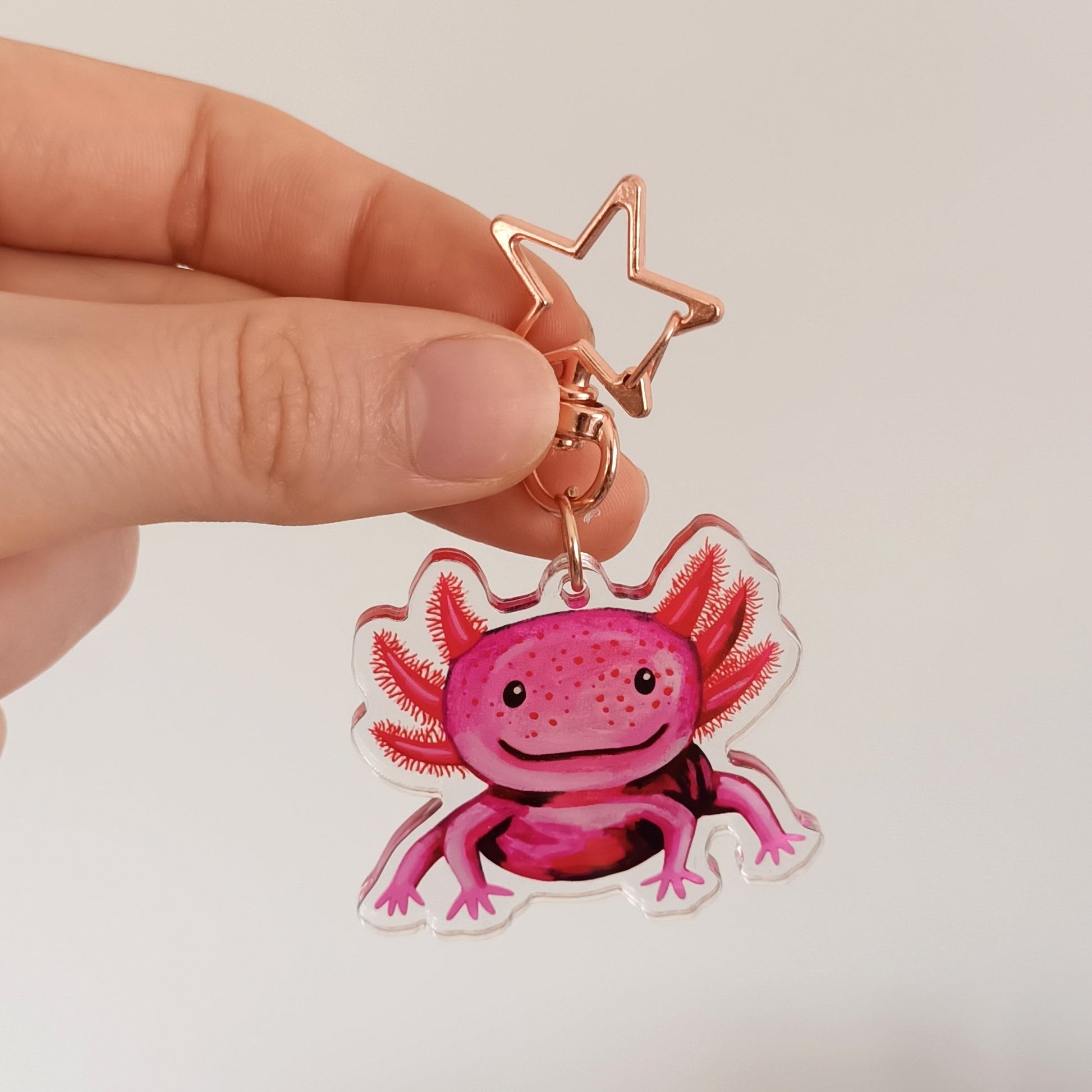 Hand holding a pink axolotl keychain with a star-shaped clip against a white wall.