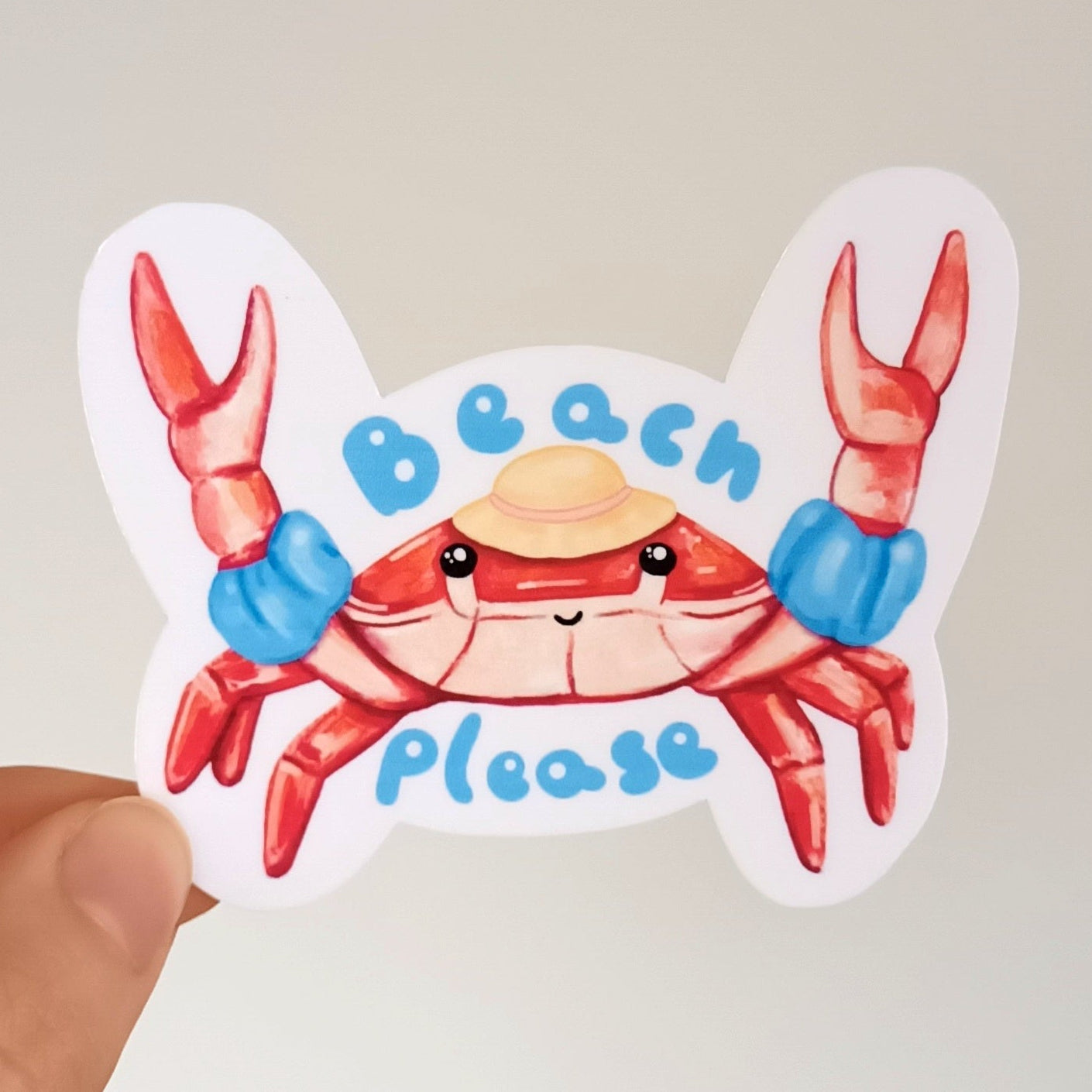 Hand holding a sticker of a cartoon crab with 'Beach Please' text against a neutral background.