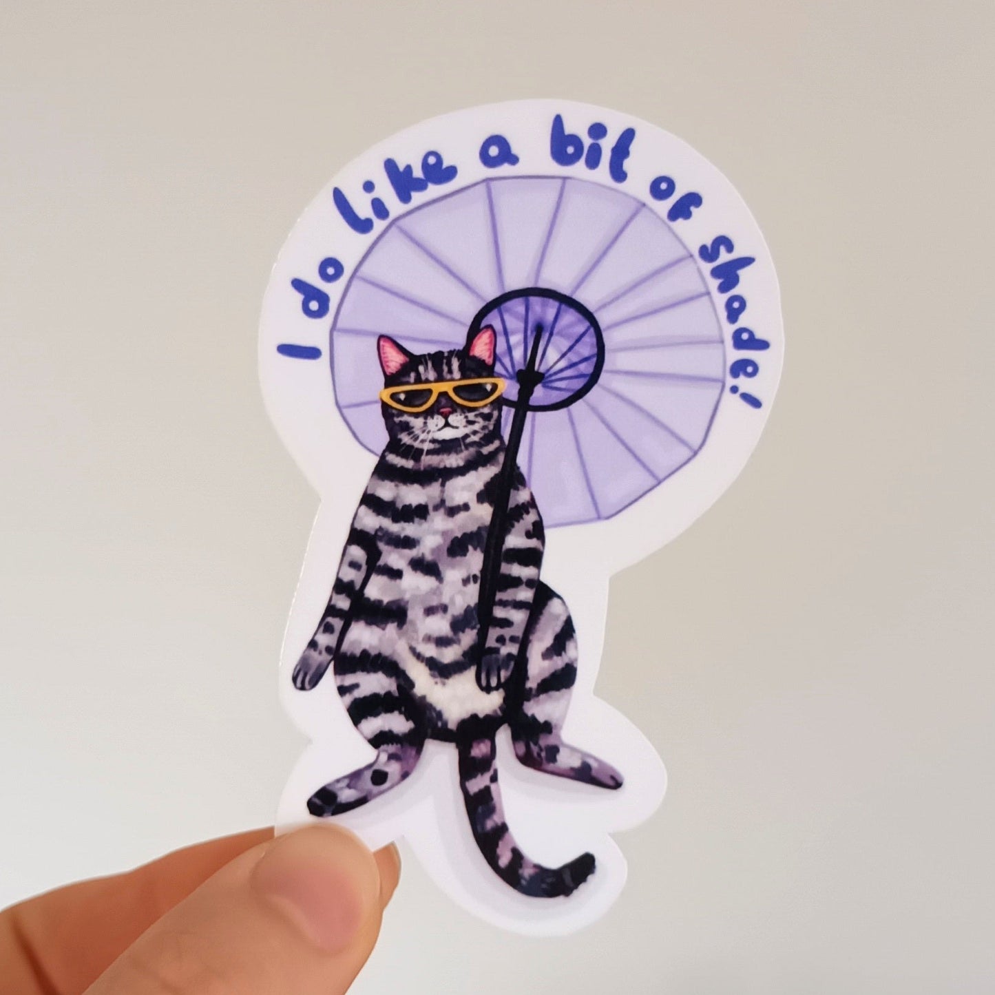 Hand holding a sticker with a cat with an umbrella design saying "I do like a bit of shade".