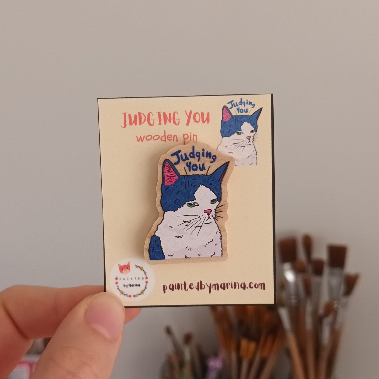 Hand holding a small box with a "Judging You" Cat Pin on a plain background