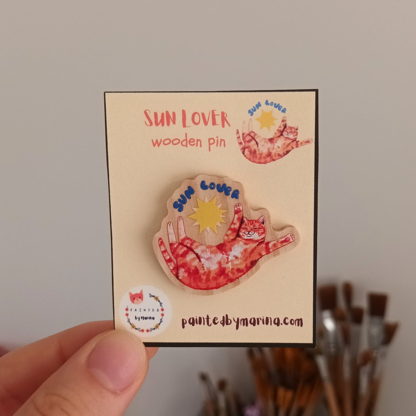 Hand holding a wooden pin with 'Sun Lover' design on a card, with art supplies in the background.