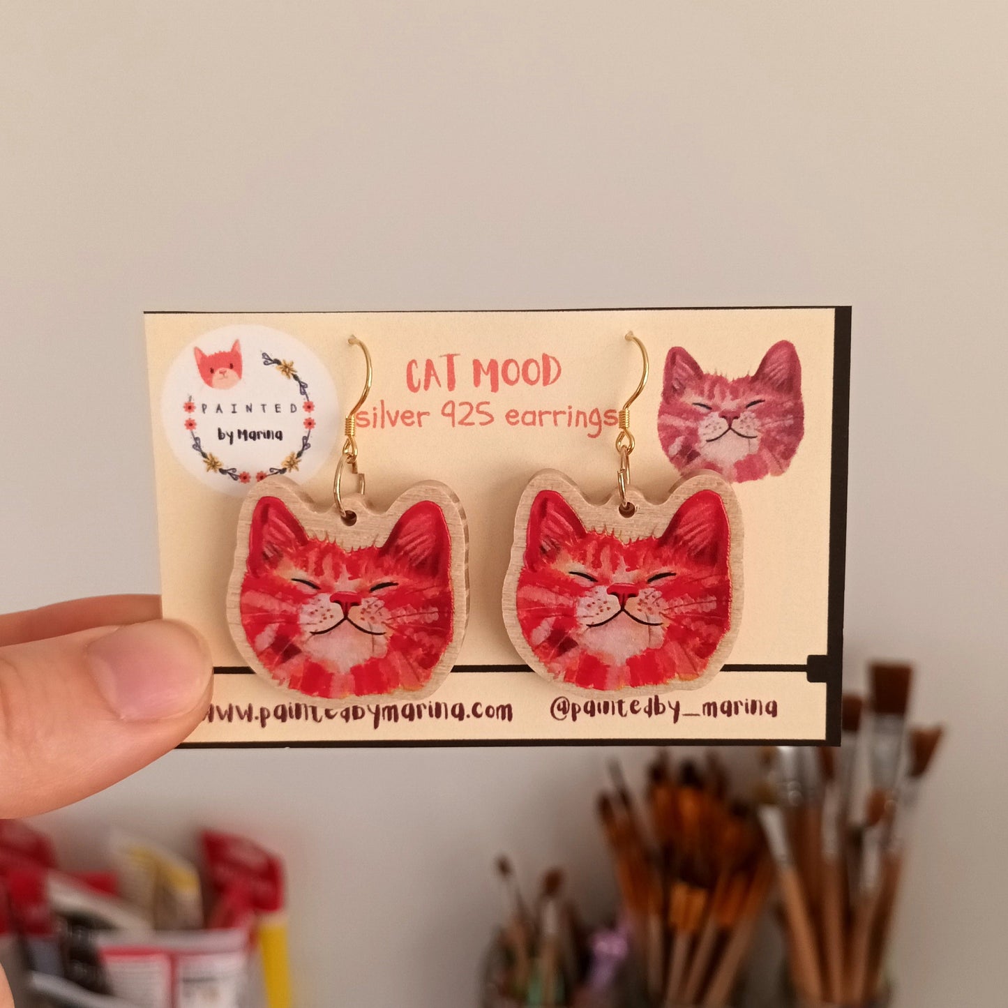 Red cat-shaped earrings on a branded card held by a hand with a blurred background of art supplies.