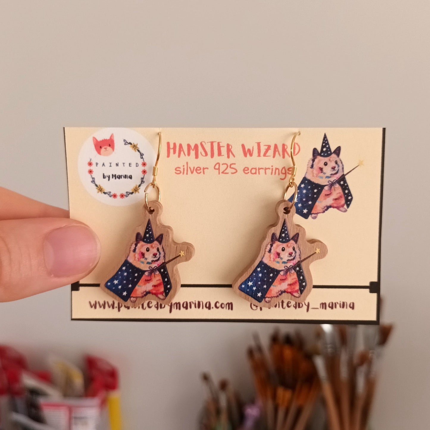 Hamster Wizard earrings in a small box held by a hand with a blurred background of art supplies.
