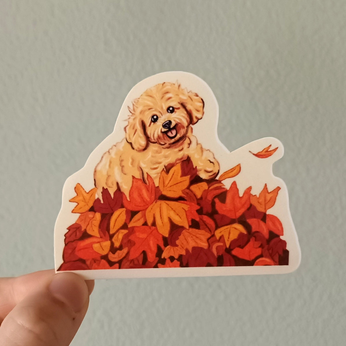 A hand holds a die-cut sticker featuring a cute cartoon puppy sitting in orange and red autumn leaves, set against a plain light gray wall.