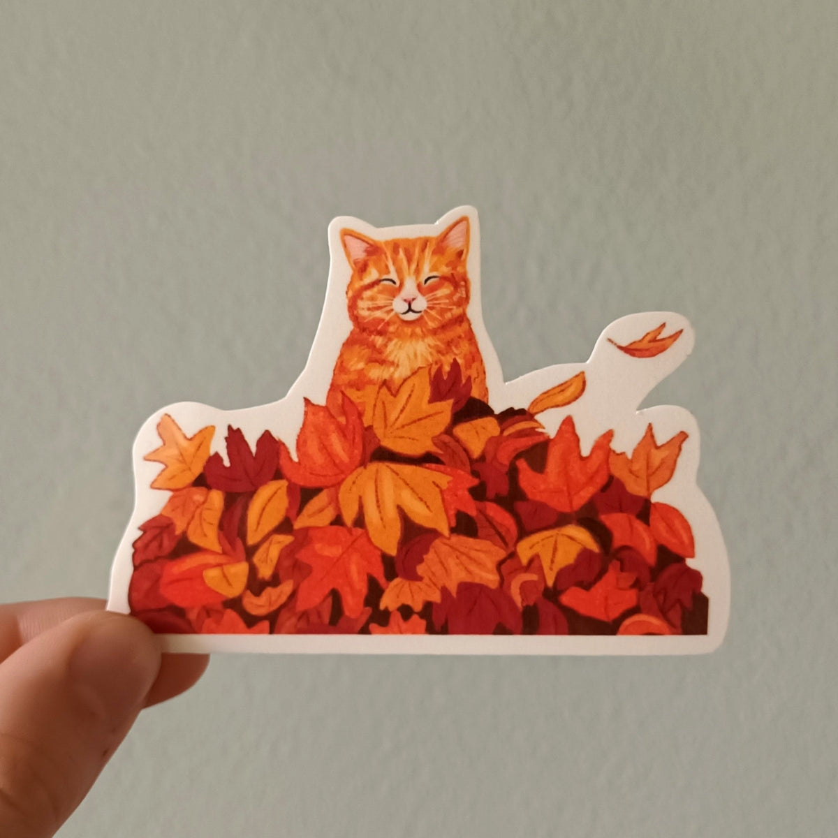 A hand holds a die-cut sticker featuring an orange tabby cat sitting on a pile of red and orange autumn leaves, against a plain light gray wall.