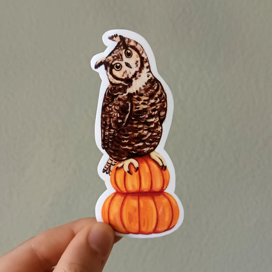 A hand holds a die-cut sticker featuring a brown owl perched on top of two orange pumpkins, against a plain light gray wall.