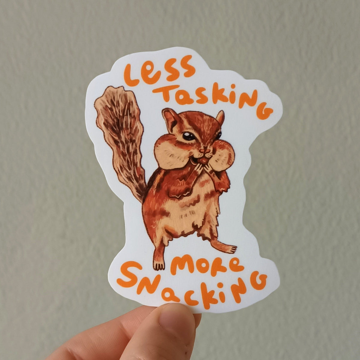 A hand holds a die-cut sticker featuring an illustrated squirrel with the text 'LESS TASKING MORE SNACKING' in orange lettering, against a plain light gray wall.