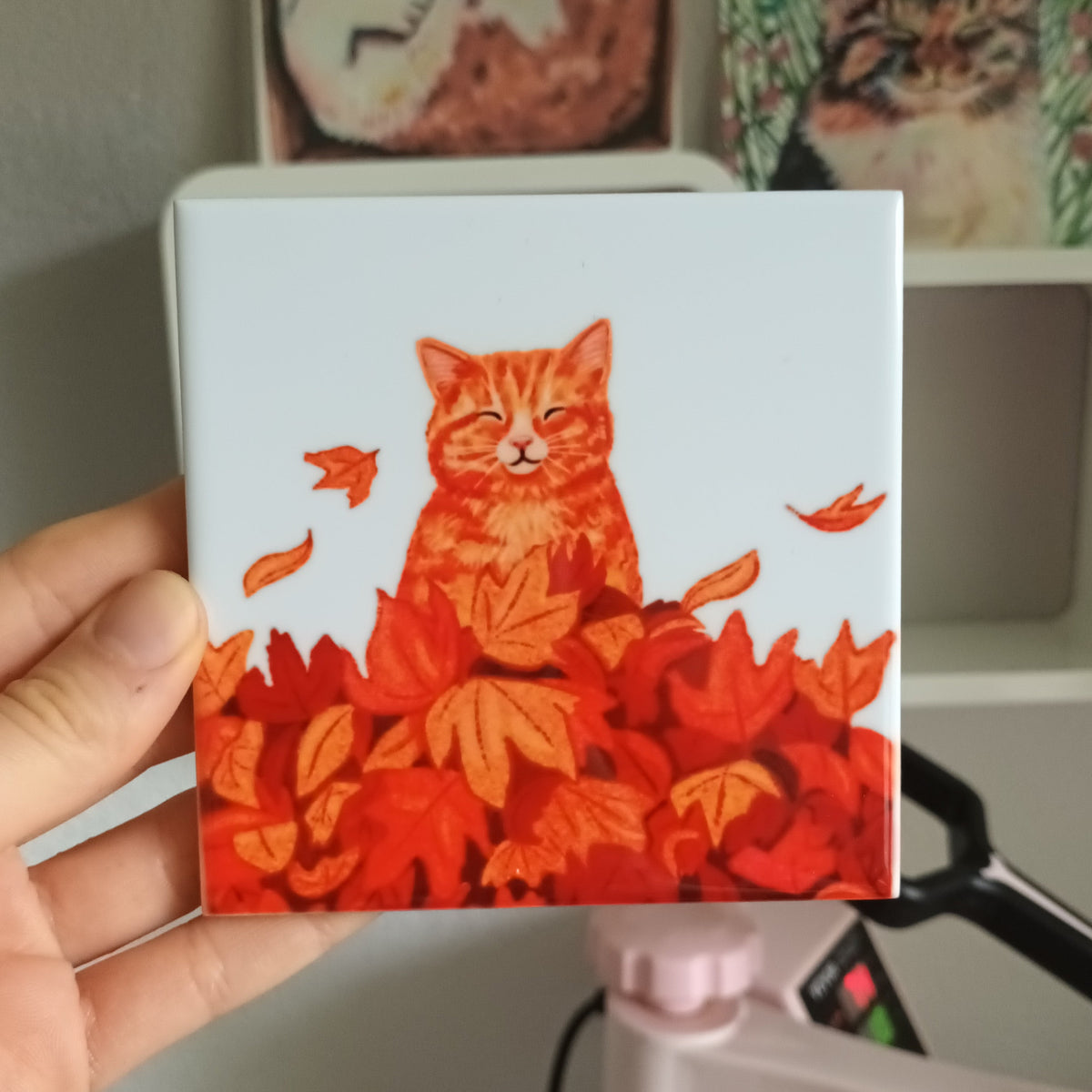A hand holds a square ceramic tile featuring an illustration of an orange tabby cat sitting on a bed of red and orange autumn leaves. In the background, a pink heat press machine and framed cat art are visible.