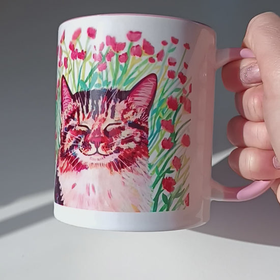 Video of handmade mug with a cute cat sitting in a meadow of pink flowers, pink handle and inside.