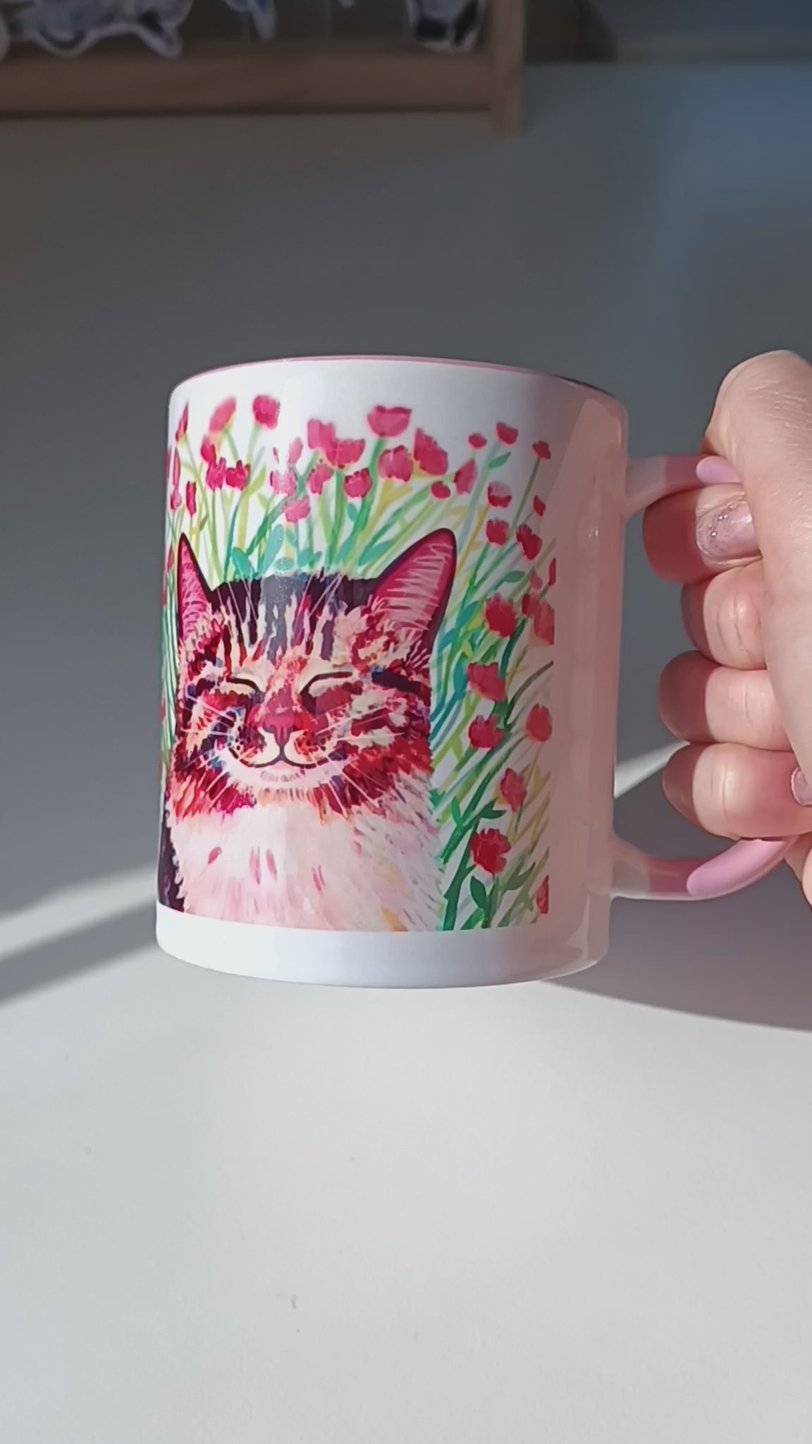 Video of handmade mug with a cute cat sitting in a meadow of pink flowers, pink handle and inside.